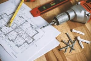 Blueprint on a wooden table with a tape measure, level bar, impact drill, and screws