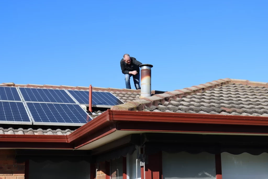 Building inspector examining tiled roof near chimney and solar panels on a sunny day in Melbourne.