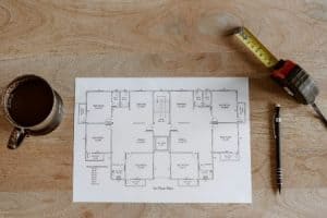 A wooden table showing a floor plan alongside a cup of coffee, retractable tape measure, and a mechanical pencil