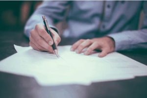 A man in casual business shirt signing on pieces of paper on a wooden table