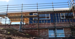 House under construction with brick lower level, timber framing above, and scaffolding around the structure.